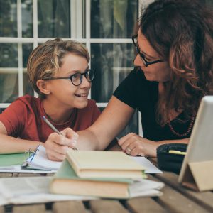 Young student doing homework at home with school books, newspaper and digital pad helped by his mother. Mum writing on the copybook teaching his son. Education, family lifestyle, homeschooling concept